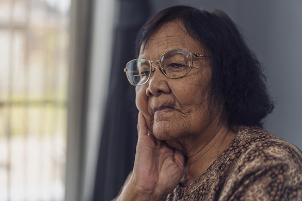 Elderly woman wearing glasses sitting inside a nursing home with her hand resting on her cheek, looking out with a thoughtful expression. (222816539)