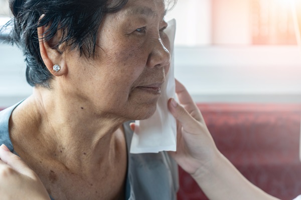 A close-up, side profile of a nursing home resident with short dark hair. A caregiver's hand is visible, gently holding a white compress to the woman's cheek to treat an injury following an assault from another resident. (1705776603)